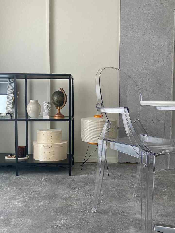 モノトーンのグレー調1LDKリビング、透明なアームチェアと黒のメタル棚、フロアランプが配置されたシンプルで無彩色の雰囲気あるインテリア