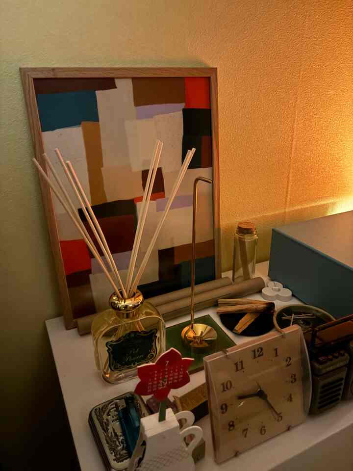 ゴールドのディフューザーと額縁が置かれた、温かみのある黄色の壁を背景にしたテーブル上のインテリア小物のある空間