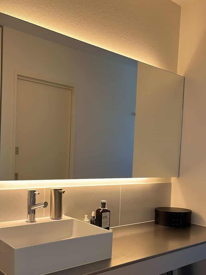 ホワイトとグレーのトーンでまとめられたシンプルモダンなバスルーム、大型の鏡と間接照明、ステンレス調の洗面カウンターが配置された印象的な空間