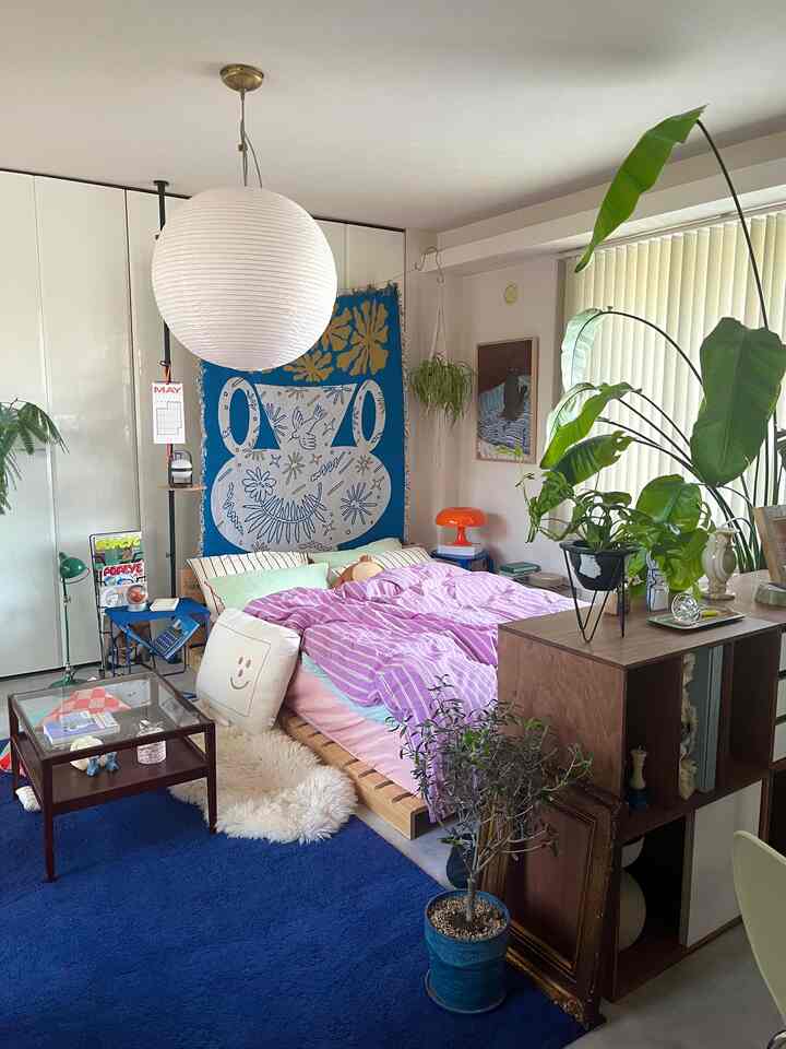 ブルーのカーペットとナチュラルウッドの家具が配置された寝室、植物とパープルのストライプ布団カバーが調和した雰囲気のあるインテリア