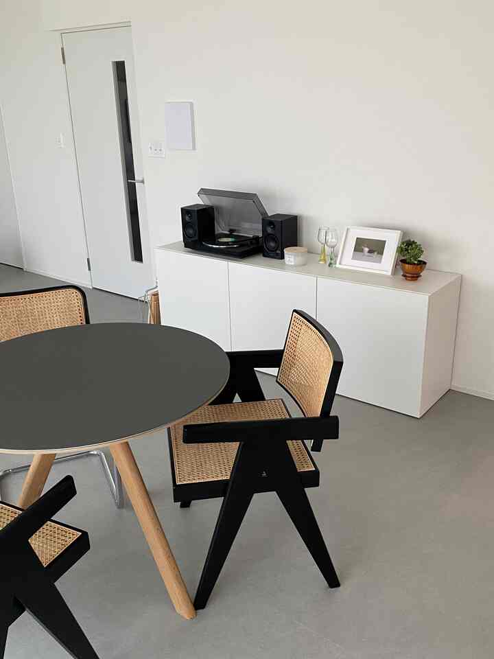 White-toned dining room featuring a round table, cane chairs, and a shelf with a record player, creating a simple and modern atmosphere