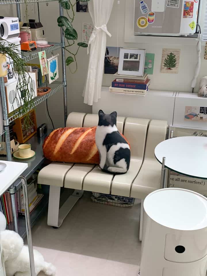 White and beige toned small room featuring cream seating with cat-shaped and bread-shaped cushions alongside a round table, creating a cozy atmosphere