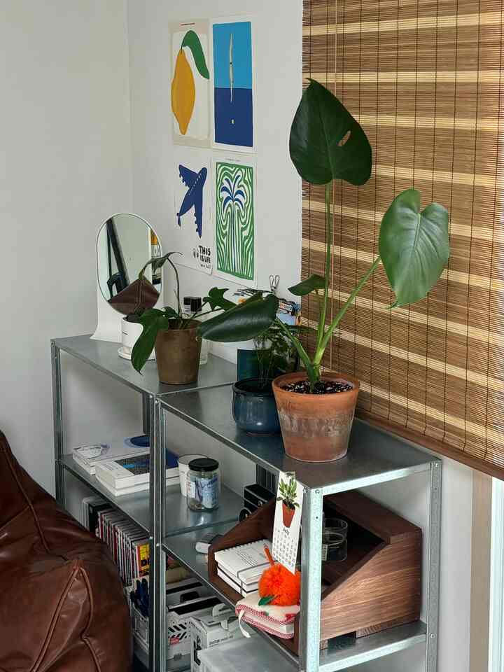 Natural-toned living room featuring metal shelves with wooden magazine rack, potted plants, and bamboo blinds creating cozy atmosphere