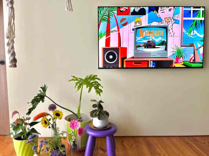 ライトブラウンの木製の床と白い壁のリビングルーム、多様な植物と紫のスツール、大きな壁掛けテレビが配置された明るい空間