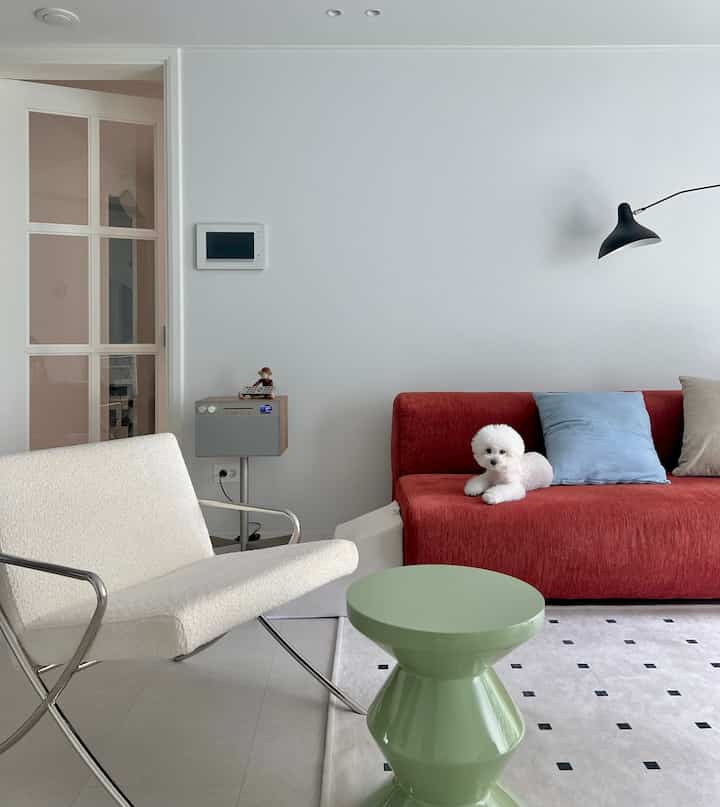 A modern living room in white and red tones featuring a red sofa, white armchair, and a green stool with clean aesthetics