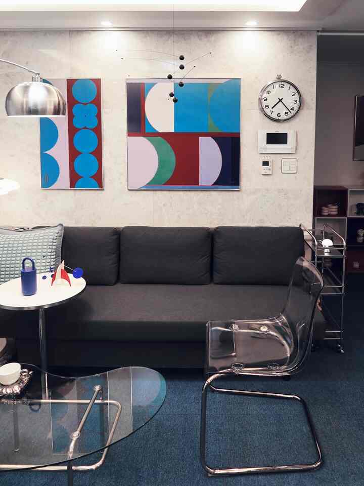 Gray-toned living room featuring a modern dark gray sofa, transparent chair, abstract art, and wall clock in a clean setting