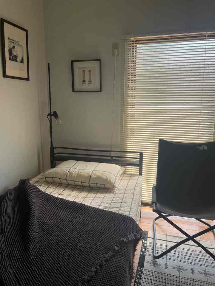 A calm blue and black toned bedroom featuring a bed with checked bedding, window blinds, and a camping chair, with a simple minimalist vibe