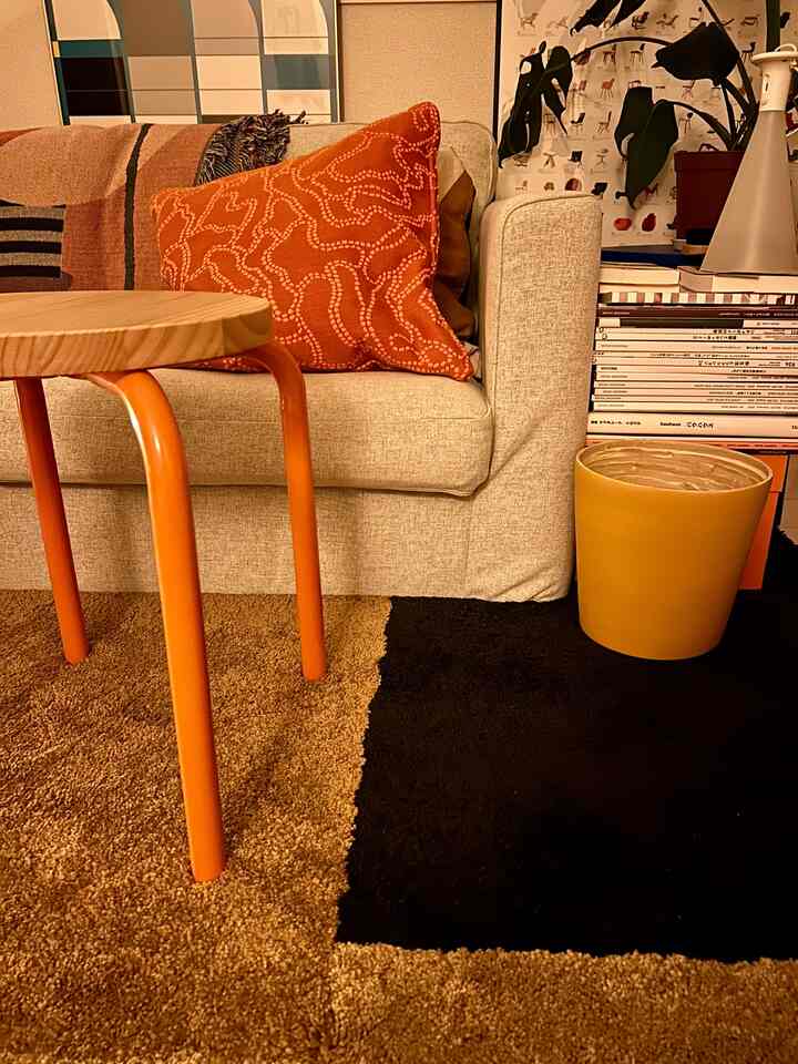 ベージュのソファとオレンジ色のスツール、ブラウンとブラックのラグが配置されたあたたかみのあるリビングスペース