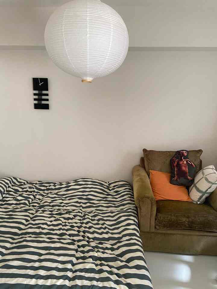 白い壁とカーキ色のソファが配置された寝室、ストライプのベッドカバーが調和するシンプルなインテリア