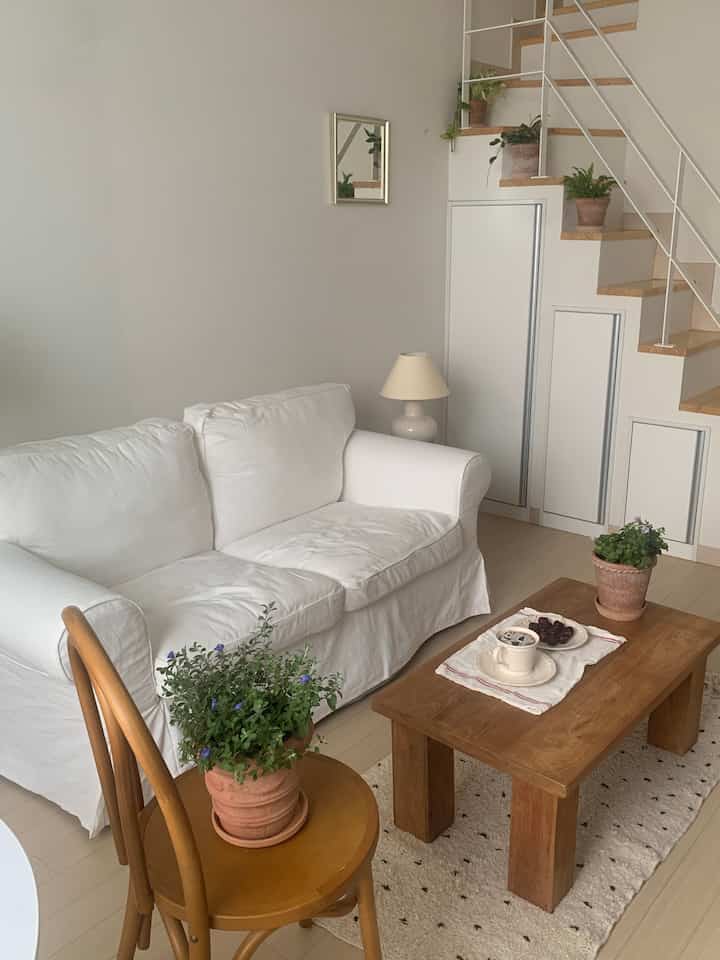 ホワイトとウッドトーンの調和が感じられるワンルームロフトのリビング、木製のソファとテーブル、植物が置かれた雰囲気のある空間