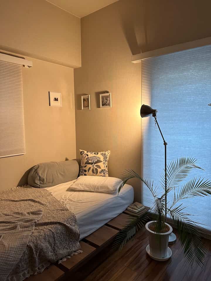 ベージュの壁と木目調の床が特徴の8畳ナチュラルな寝室、木製ベッドフレームと白い寝具、そしてフロアランプと植物が配置された印象的な空間