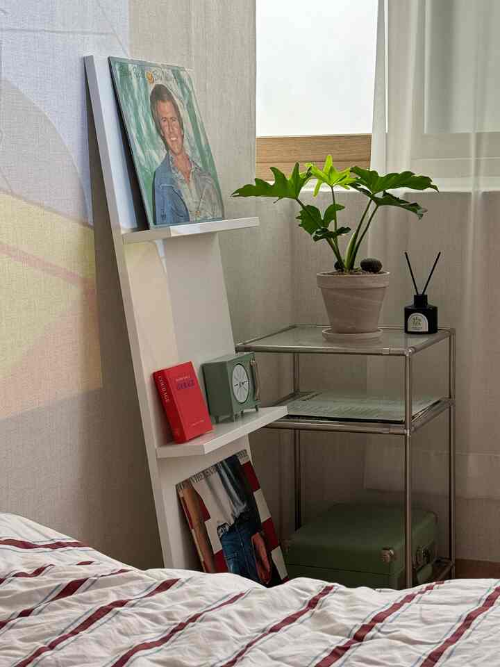 柔らかな白色の寝室コーナー、白い棚と金属のナイトテーブルの上に置かれた観葉植物が配されたシンプルな空間