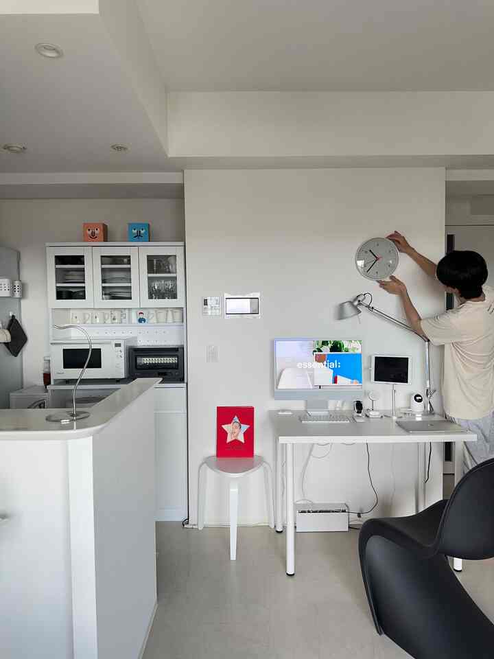 Modern white-toned home office space featuring a simple desk with clock and kitchen area harmoniously arranged