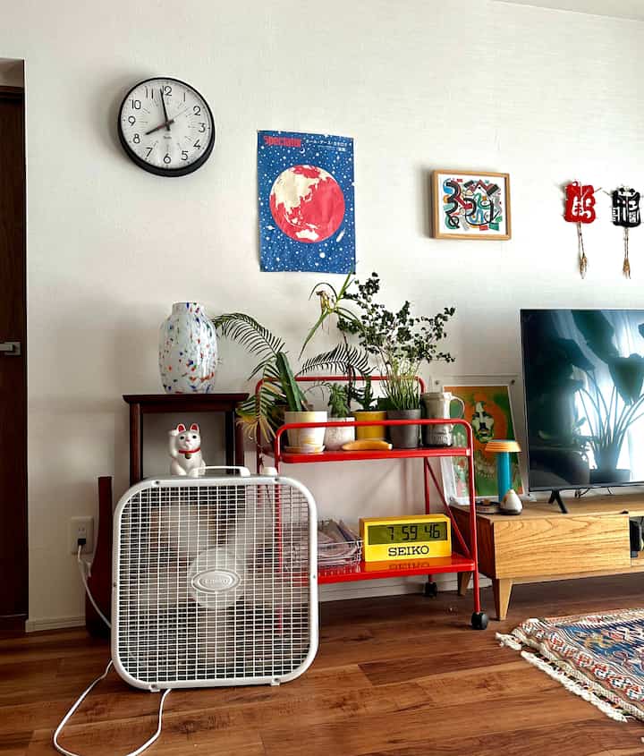 白い壁と木目調の床のリビング、時計、ボックスファン、植物、テレビ台が配置された自然な雰囲気のインテリア