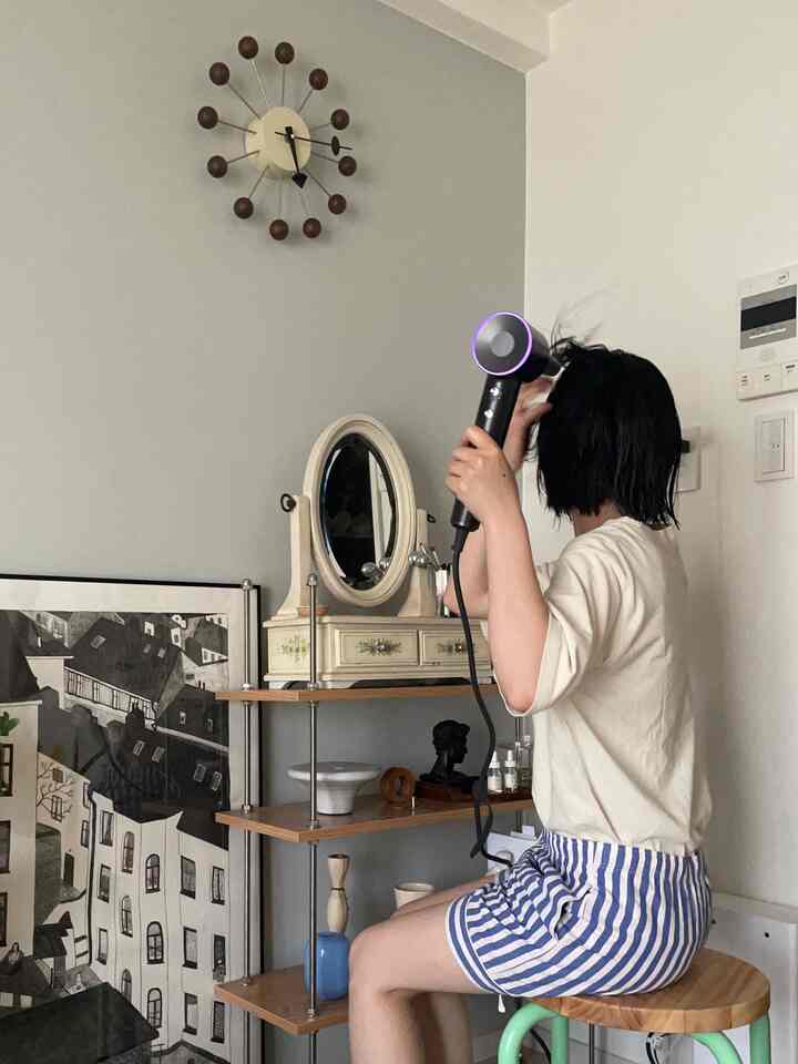 Natural modern studio apartment in white and gray tones, featuring a wooden stool, wall clock, art print in a dressing area where person is drying hair