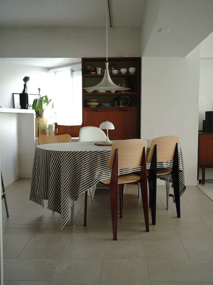 白い壁と木目調家具が調和するダイニング、中央にストライプのテーブルクロスをかけたテーブルと多様な椅子が配された、自然でモダンな雰囲気の空間