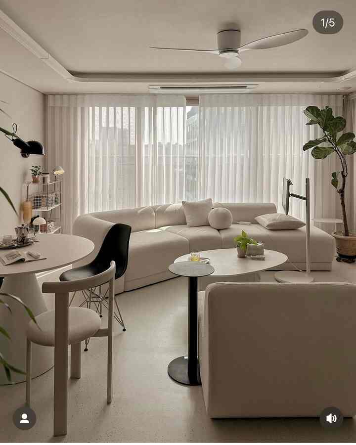 Modern living room in white tones featuring a large curved sofa, coffee tables, and a tall plant in a bright serene space