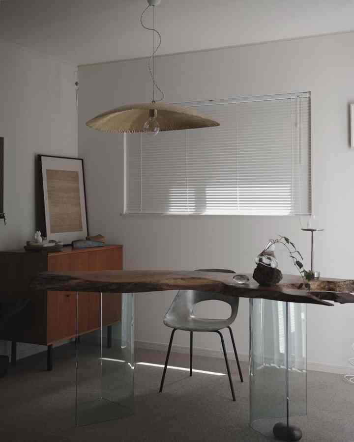 Modern dining room with white walls and blinds, brown wood slab table with glass legs, and a central brass pendant light