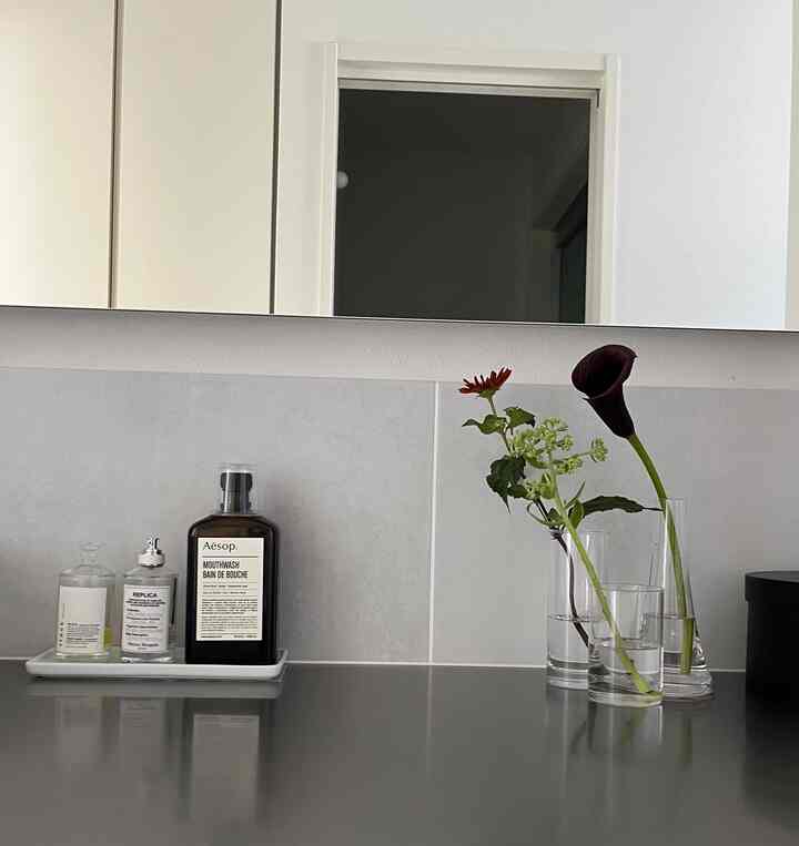 グレーのタイルとステンレスのシンクが特徴の白を基調としたバスルーム、ガラスの花瓶に活けられた新鮮な花が配置された清潔感のある空間