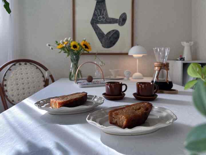 白いテーブルクロスが敷かれたダイニングルーム、コーヒーとハニートースト、ひまわりの花瓶、自然光と温かみのあるテーブルランプが配置された雰囲気あるインテリア