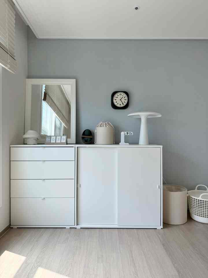 淡いグレーの壁と白い家具が調和したシンプルな収納スペース、時計とテーブルランプが飾られた清潔感のあるインテリア