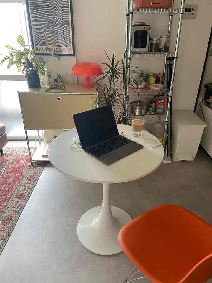 White and orange toned 1DK living space featuring round dining table, orange chair, open shelves and coffee machine creating cozy remote work environment