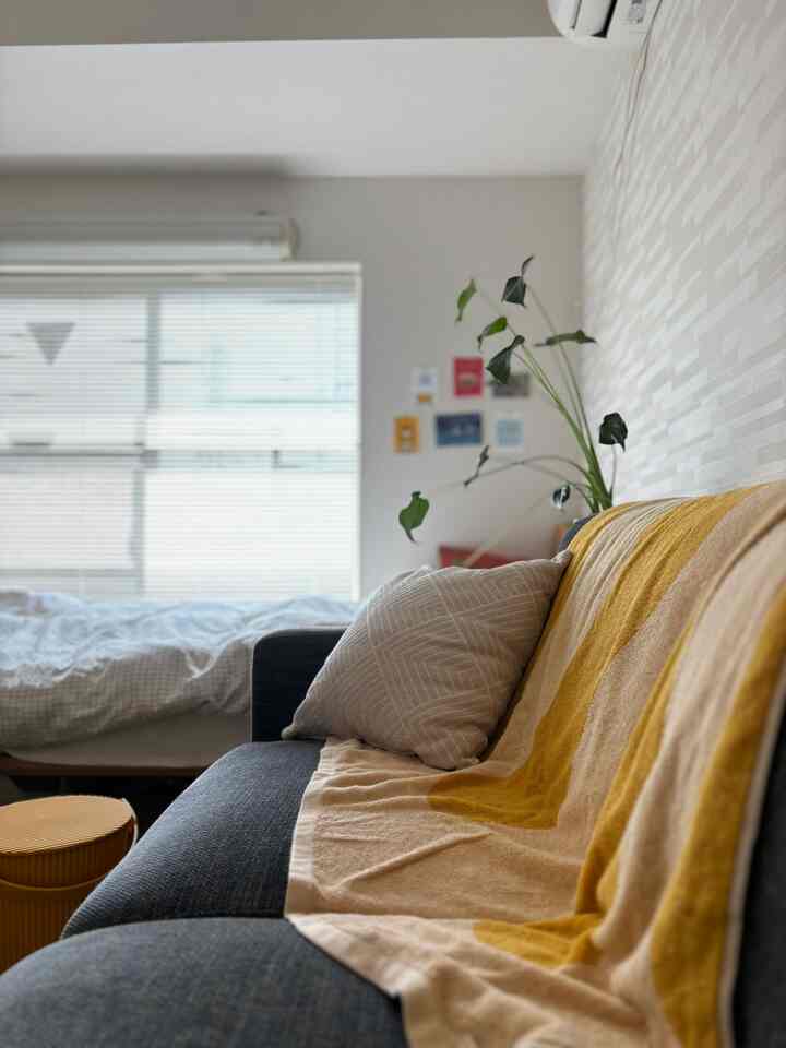 明るくナチュラルな色調の1Kの一人暮らし寝室、黄色のブランケットがかかったダークグレーのソファと明るい窓のブラインドがある空間