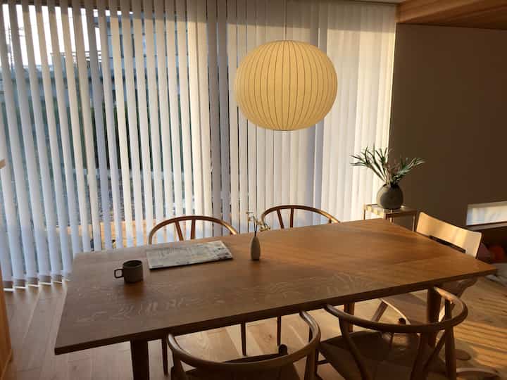 明るくナチュラルトーンのダイニング、木製のテーブルと椅子、天井に吊るされたバブルペンダント照明とバーチカルブラインドが配置されたシンプルな空間