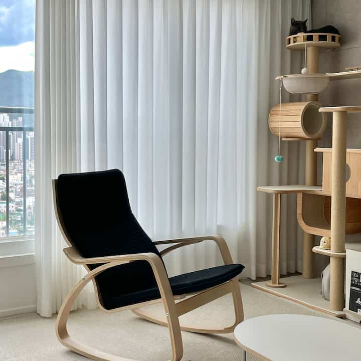 明るいナチュラルトーンのリビング、白いカーテンと黒いアームチェア、猫がいるキャットタワーが配置された居心地の良い空間