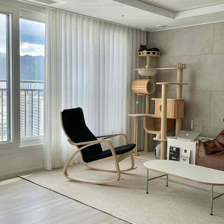 ナチュラルな色調のリビング、中央に黒いクッションの木製ロッキングチェア、ベージュのラグ、右側にキャットタワーとビーンバッグが配置された温かみのあるインテリア