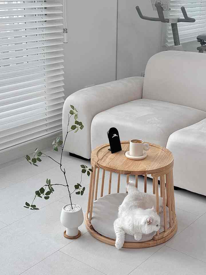 白と木目調が調和したリビング、木製ペットハウス兼コーヒーテーブルとクッション、白い猫がくつろぐ雰囲気のあるインテリア