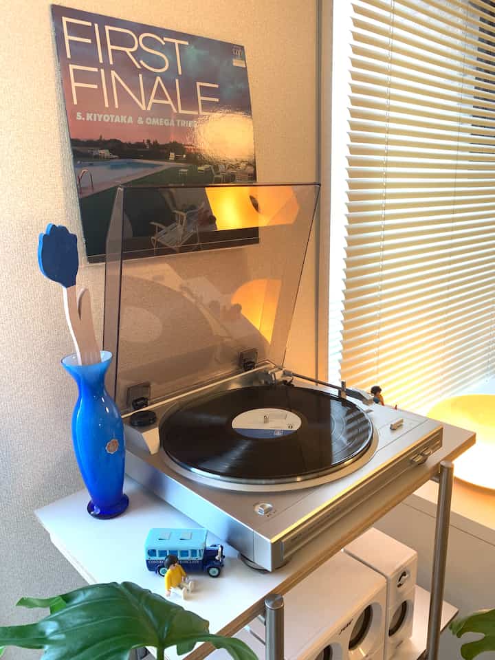 白い棚に置かれたレトロなデノンのターンテーブルと青い花瓶、窓にかかるブラインドが調和する音楽空間