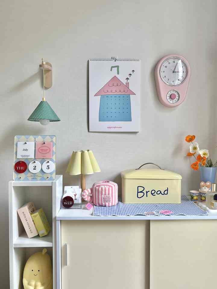 パステルカラーの白とベージュのキッチンスペース、黄色いパンケースとピンクの壁時計が調和した清潔感のあるインテリア