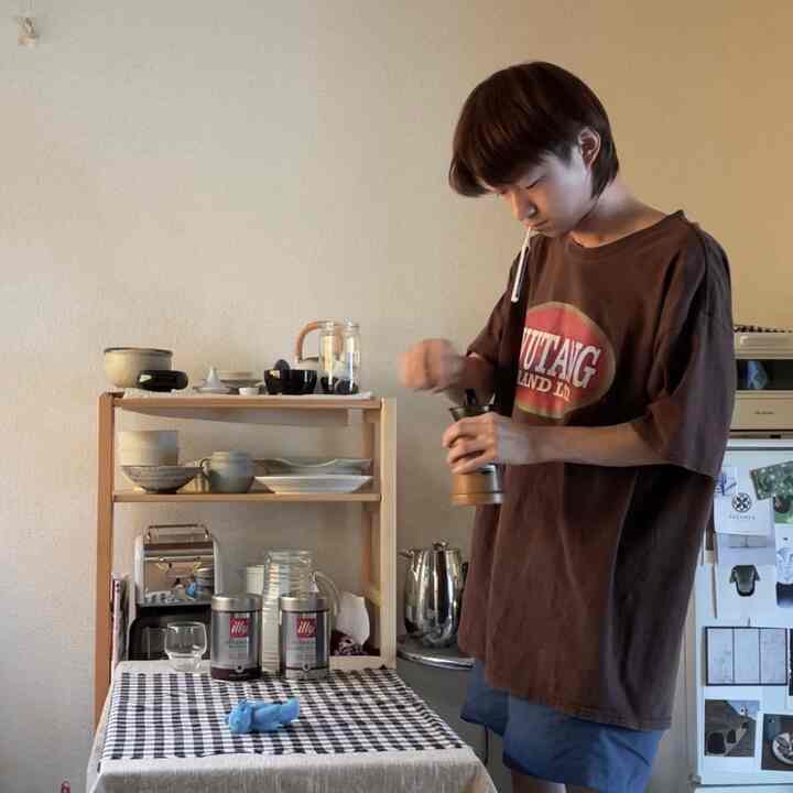 ブラウンの壁とダイニングテーブルがあるキッチンで、人物が手動コーヒーグラインダーを使うモダンなおうちカフェの雰囲気あるインテリア