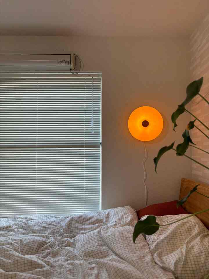 白とナチュラル色調の1K寝室、ドーナツ型のオレンジ色の壁ランプとパターン寝具が配置された雰囲気のあるインテリア