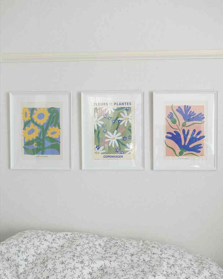 白い壁にボタニカルアートのポスターが3枚掛かっており、下に柄のある白い掛け布団がある寝室のインテリア
