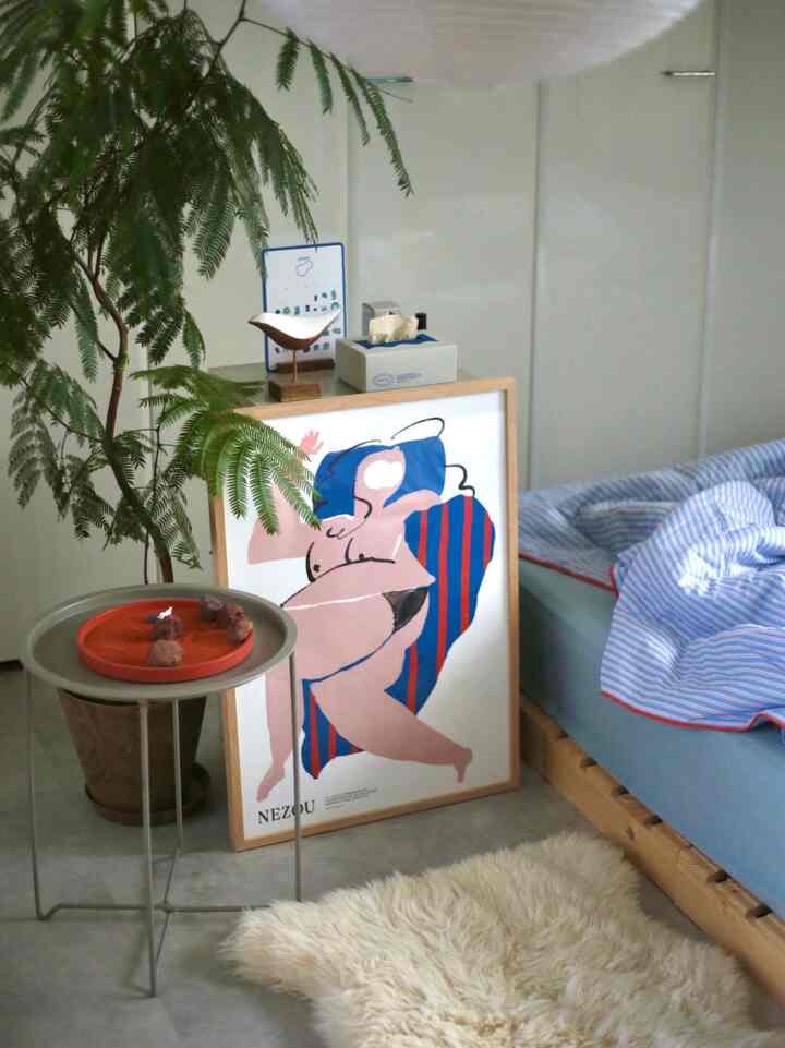 白い壁と青い寝具のナチュラルな寝室、木製ベッドフレームと観葉植物、ポスターが落ち着いた雰囲気を演出しています