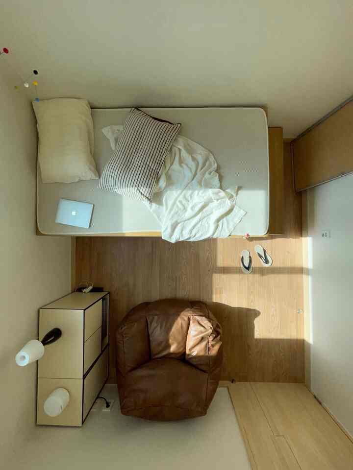 明るいベージュの壁と木目調の床があるコンパクトな一人暮らし用寝室、ブラウンのビーンバッグとベッドが配置された雰囲気のあるインテリア