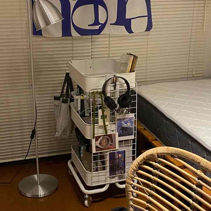 白い寝具と茶色の木目調の床、ナイトテーブル代わりのトロリーとブラインドが配置された寝室の落ち着いたインテリア