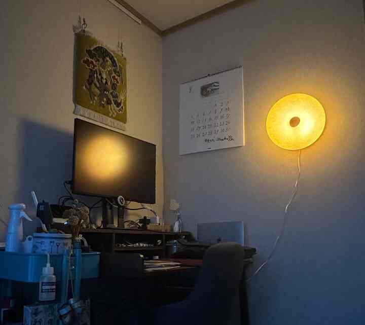 ベージュと白の色調の狭いワンルーム、壁にドーナツ型の照明が灯り、デスクとモニターが配置された落ち着いたホームオフィス空間