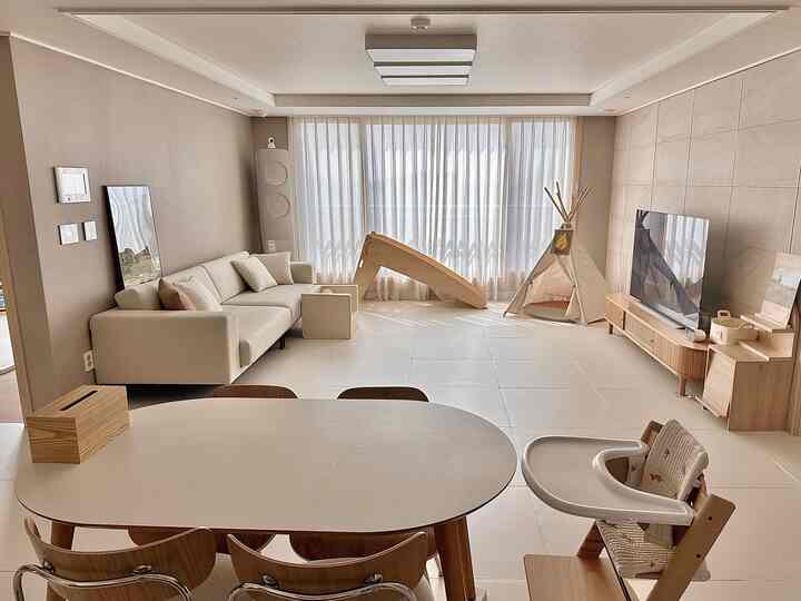 ナチュラルベージュの広々としたリビング、木製ダイニングテーブルと子供の遊具、カーテンが配置された明るく温かみのあるインテリア