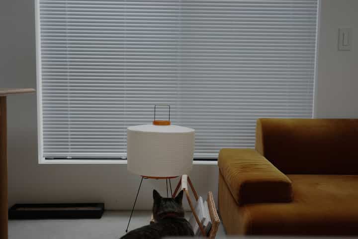 白いブラインドとブラウンソファ、AKARIのフロアランプ、猫の爪とぎが配されたナチュラルな雰囲気のリビング