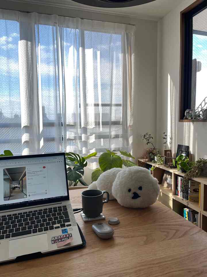 白と木目調の自然な雰囲気のホームオフィス、窓際のダイニングテーブルにノートパソコンと犬の形のクッションが配置された印象的な空間