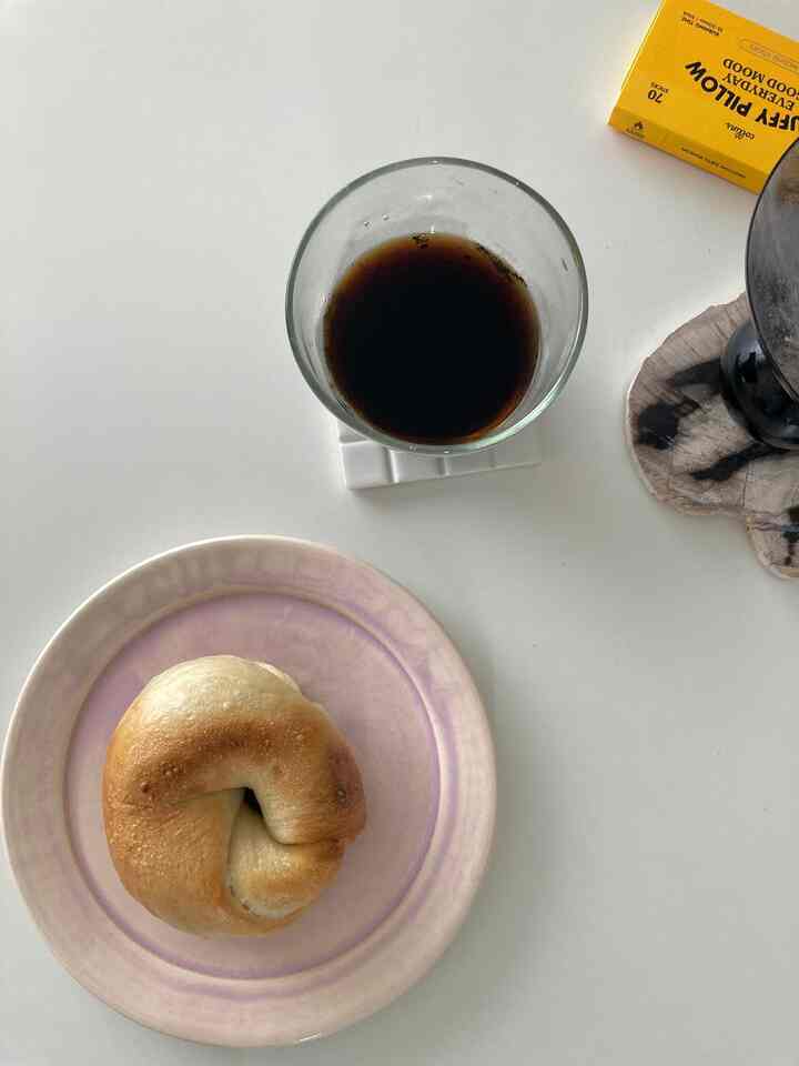 White toned table featuring a glass of black coffee on a coaster and a bagel on a plate for a simple breakfast setup