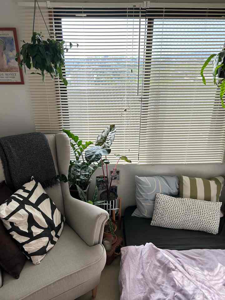 A bedroom with white blinds, natural color armchair, and green plants creating a cozy atmosphere
