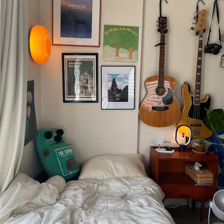 暖かいオレンジ色の壁照明と木製家具があるミッドセンチュリーモダンスタイルの8畳の寝室、壁にギターと絵画が配置された印象的な空間