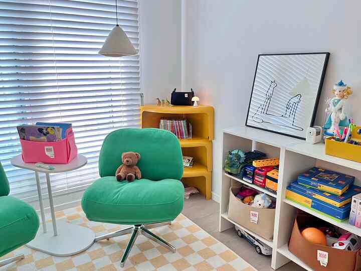 明るいホワイトトーンの子供部屋、緑のファブリックアームチェアと黄色のコーナーシェルフ、おもちゃの棚が配置された雰囲気のある読書スペース