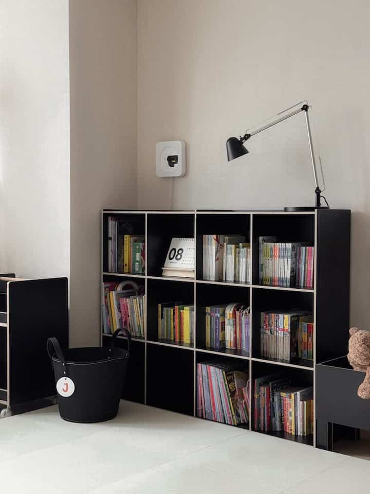A clean kids' room space featuring a black bookshelf with books, a desk calendar, and a table lamp against white floor and walls