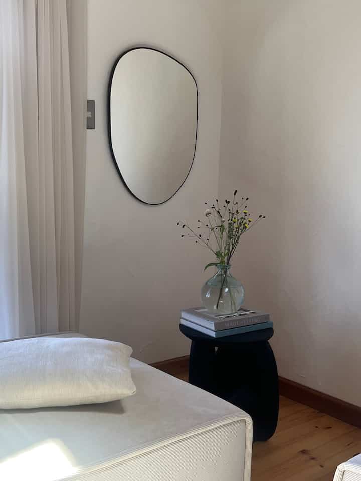 白い壁と木目調の床が特徴の北欧風寝室、黒のサイドテーブルと丸い鏡が配置された自然でシンプルなインテリア