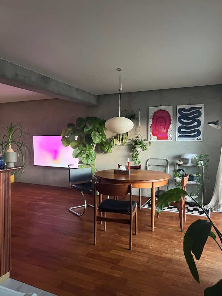 A dining room featuring wood tones and gray walls with a round dining table, pendant light, colorful posters, and plants creating a cozy atmosphere